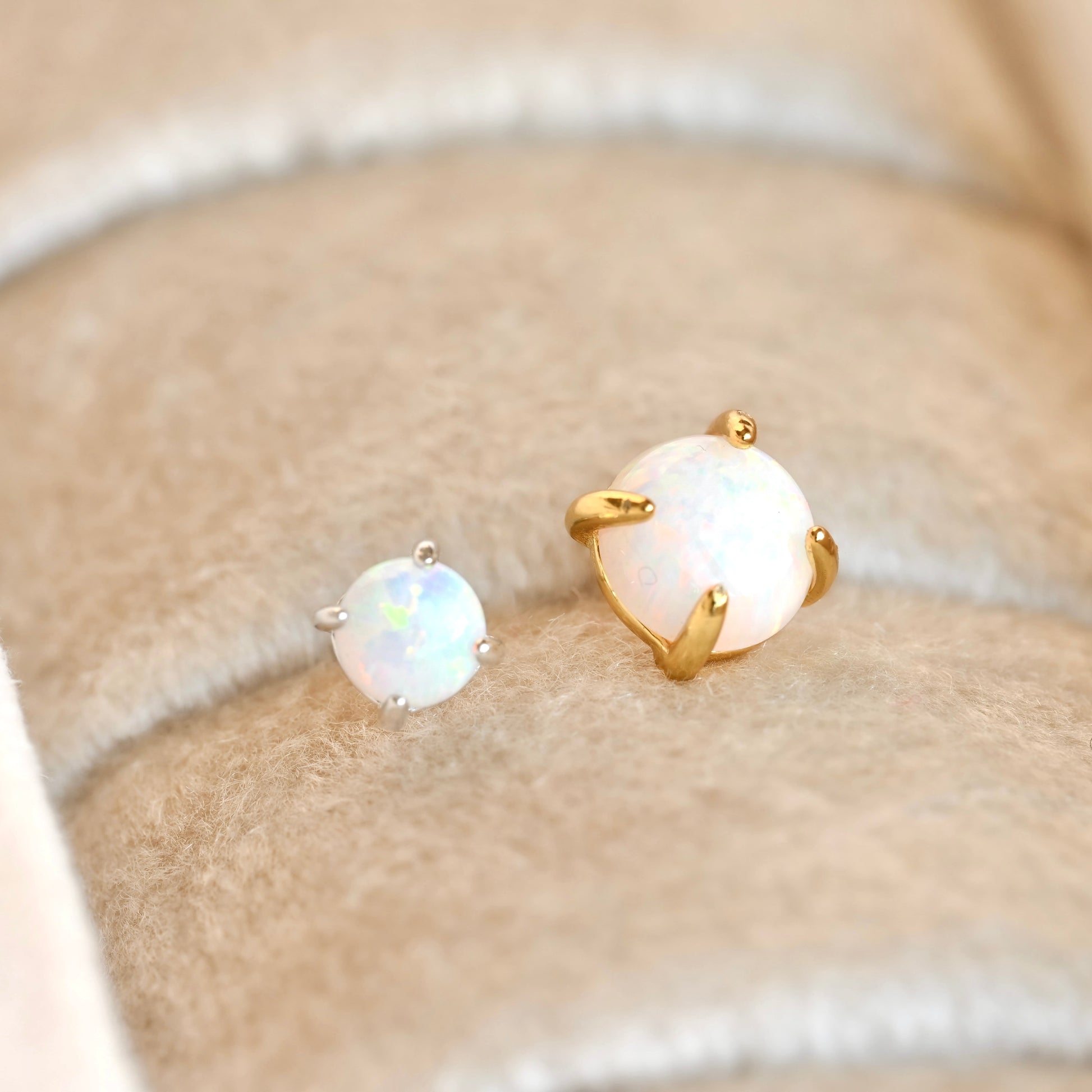 A pair of stud earrings with round white opal gemstones in gold and silver settings, displayed on a beige felt jewelry ring cushion.