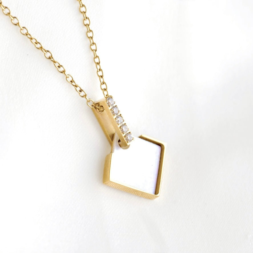 A gold chain necklace with a geometric pendant featuring a white inlay and a vertical row of clear square-cut stones, displayed on a white textured fabric background.