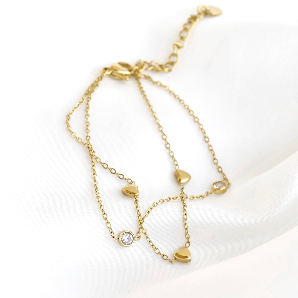 A ddouble-layered gold chain bracelet featuring multiple small gold heart charms, one of which is set with a clear round gemstone, displayed on a soft white fabric background.