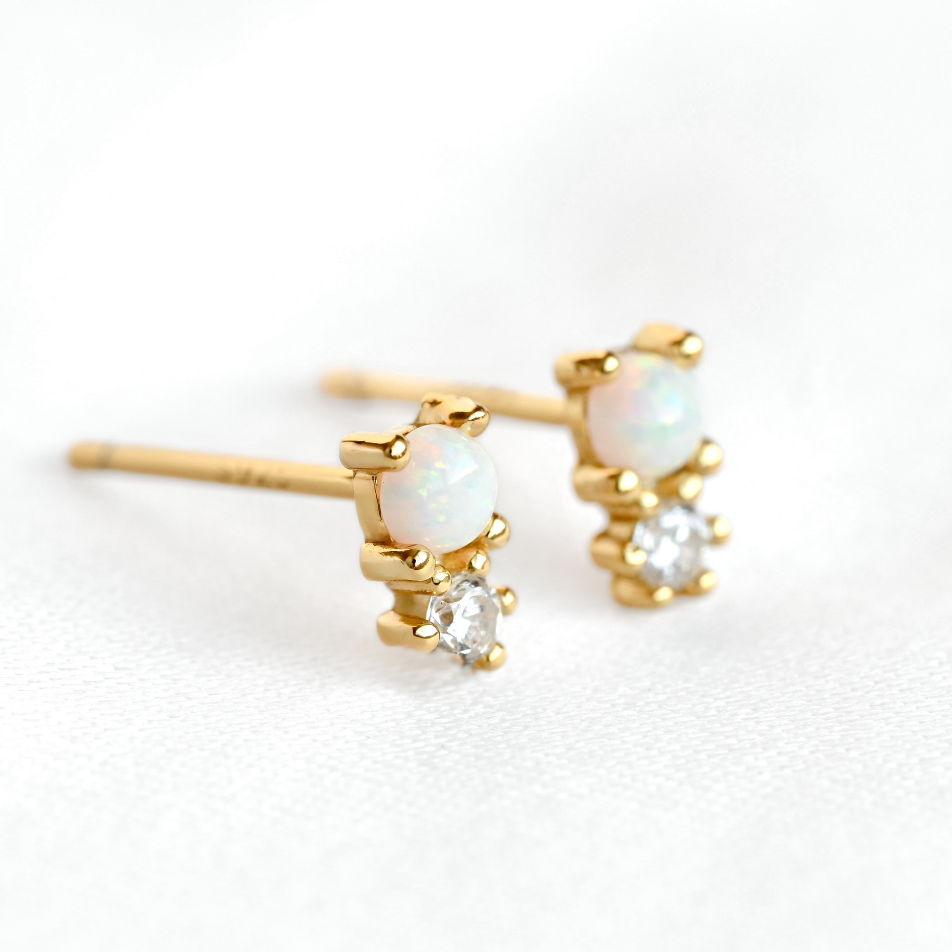 A pair of gold stud earrings featuring oval white opal and round clear crystal settings, photographed on a clean white background.