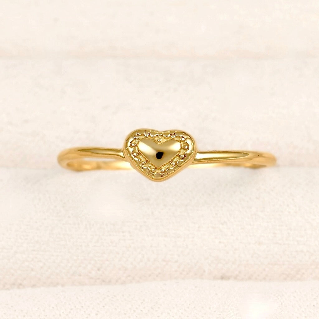 A delicate gold ring featuring a small heart-shaped centerpiece with a textured, beaded border and a polished center, displayed on a soft, textured cream fabric background.