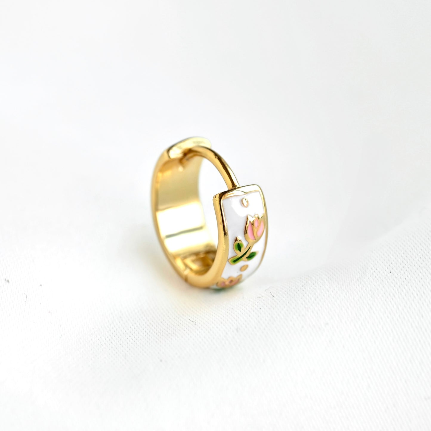A gold-plated ring with a white enamel inlay featuring a small pink tulip flower and green leaves, displayed on a white textured fabric background.