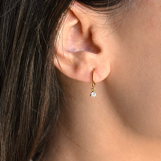 Close-up of an ear wearing gold huggie earrings with a clear gemstone.