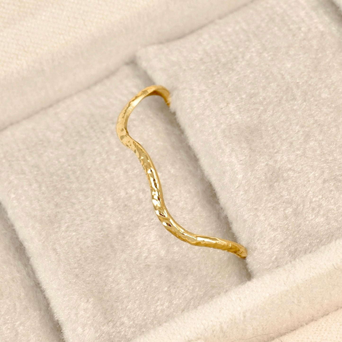 A textured gold ring with a wavy, organic shape resting on a soft beige velvet jewelry display.
