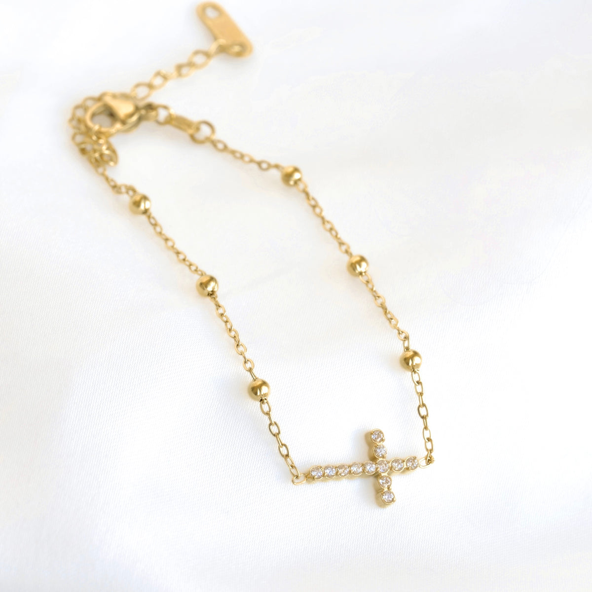 A delicate gold chain bracelet featuring a small cross charm paved with clear stones, displayed on a soft white fabric background.