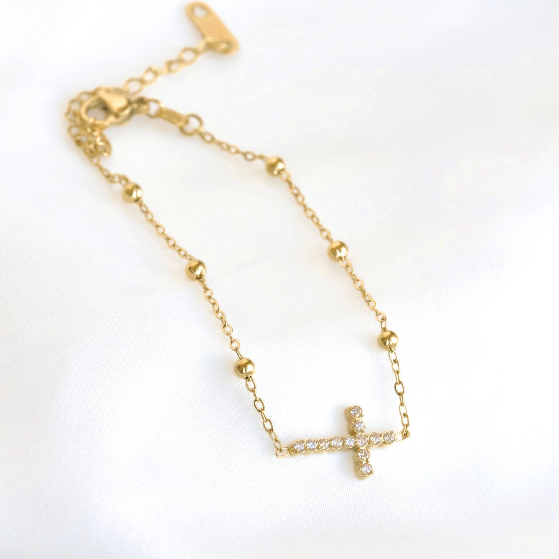 A delicate gold chain bracelet featuring a small cross charm paved with clear stones, displayed on a soft white fabric background.