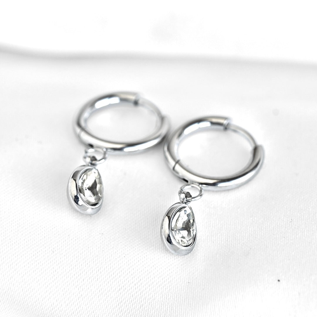 A pair of silver huggie hoop earrings with dangling teardrop clear rhinestone charms, displayed on a white textured fabric background.
