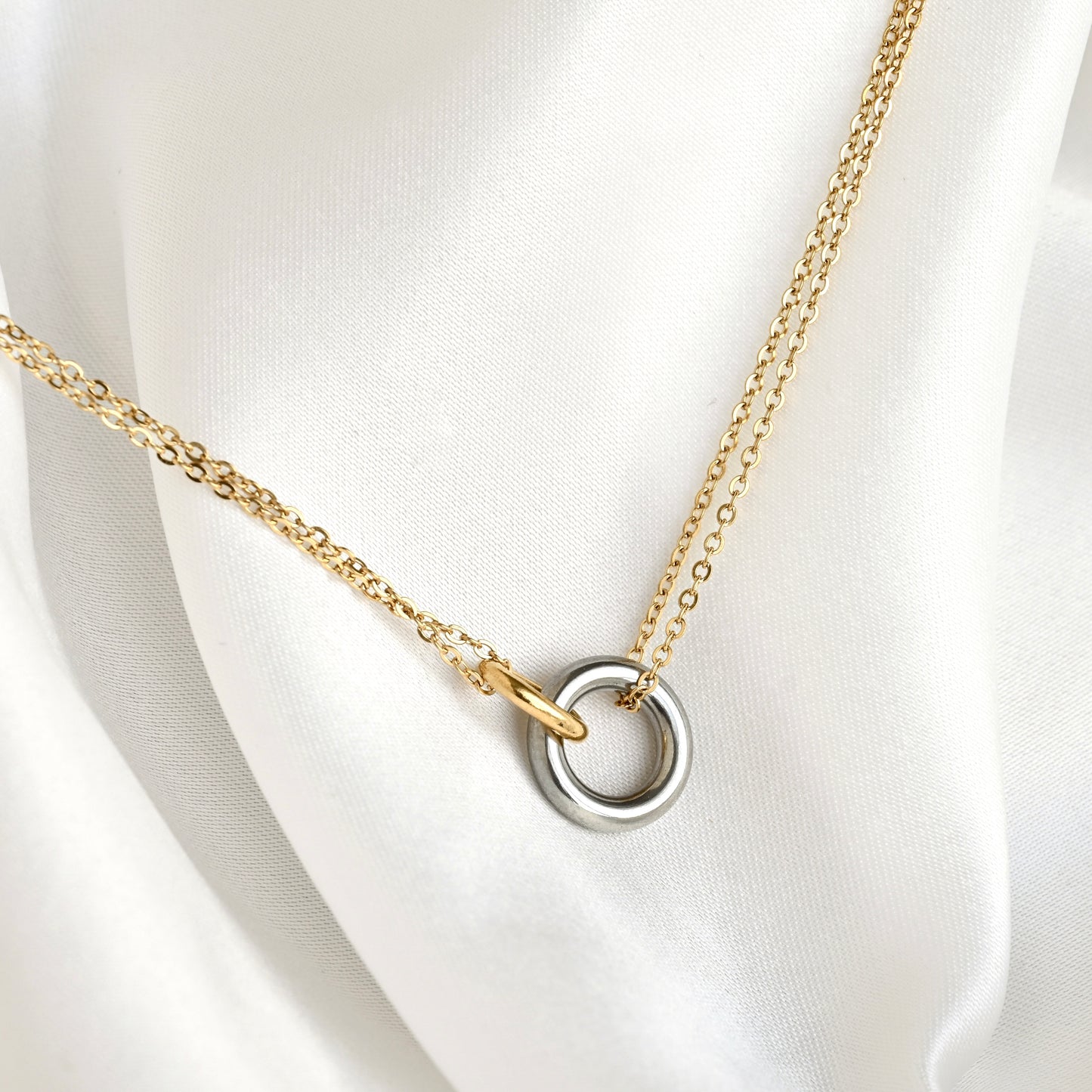 A delicate gold chain necklace with a two-tone interlocking ring pendant, featuring one matte silver ring and one polished gold ring, displayed on a draped white satin fabric.
