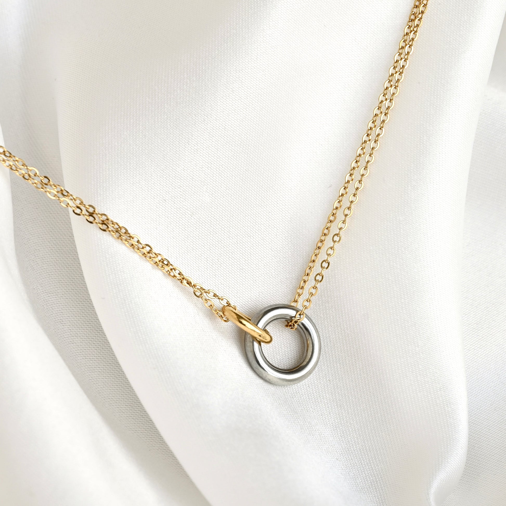 A delicate gold chain necklace with a two-tone interlocking ring pendant, featuring one matte silver ring and one polished gold ring, displayed on a draped white satin fabric.