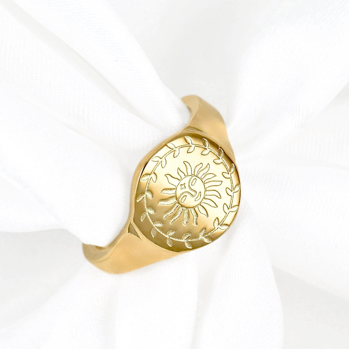 A gold signet-style ring with an engraved sun and leaf motif, displayed on a white fabric napkin.