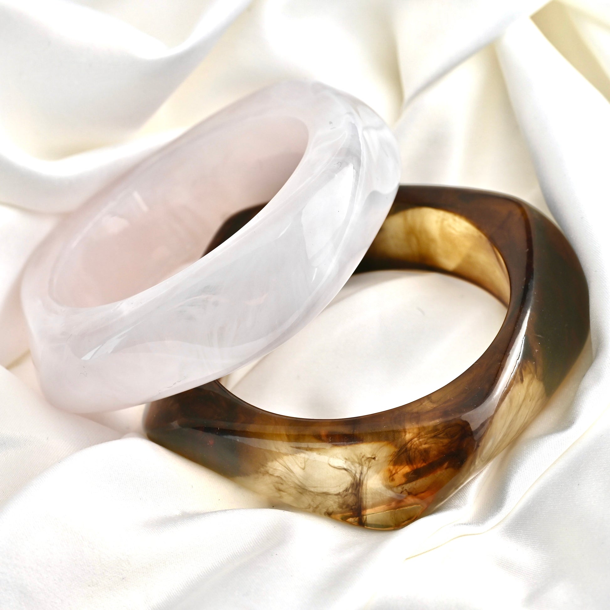 Two angular bangles displayed on draped white fabric: one translucent white and one translucent dark brown with amber-gold marbling.