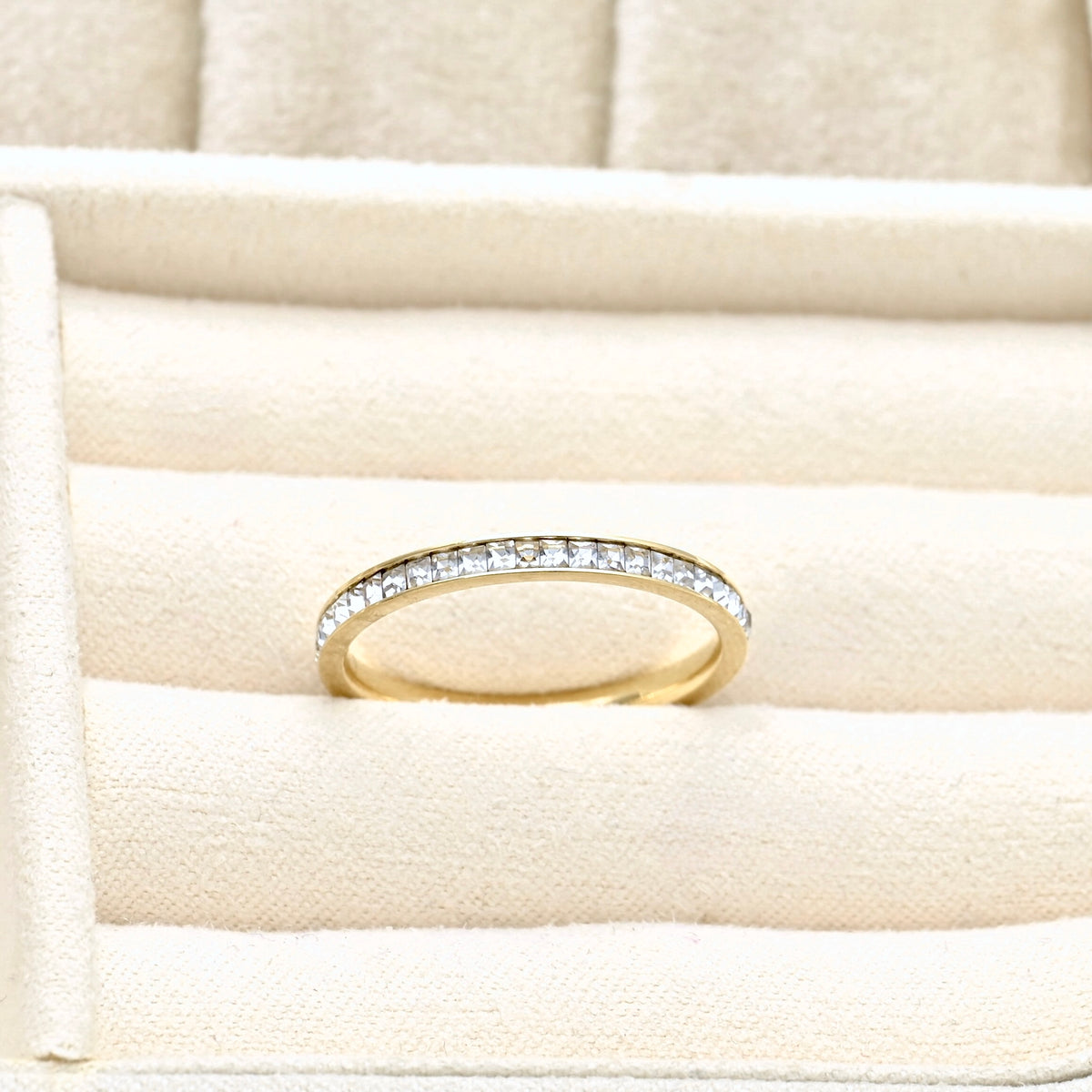 Front top view of a thin gold eternity diamond ring with channel set squre cubic zirconia stones in a beige jewelry box