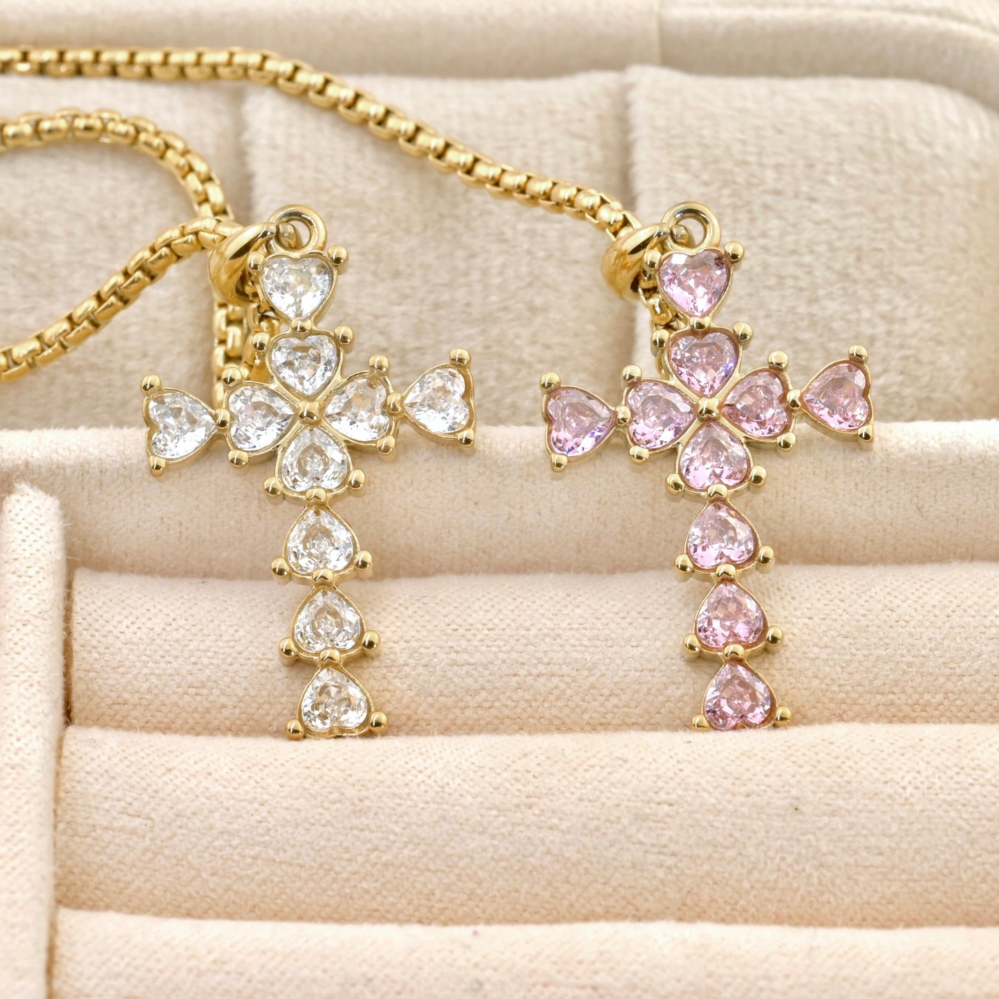 Gold cross pendants with clear and pink heart-cut stones on a beige surface
