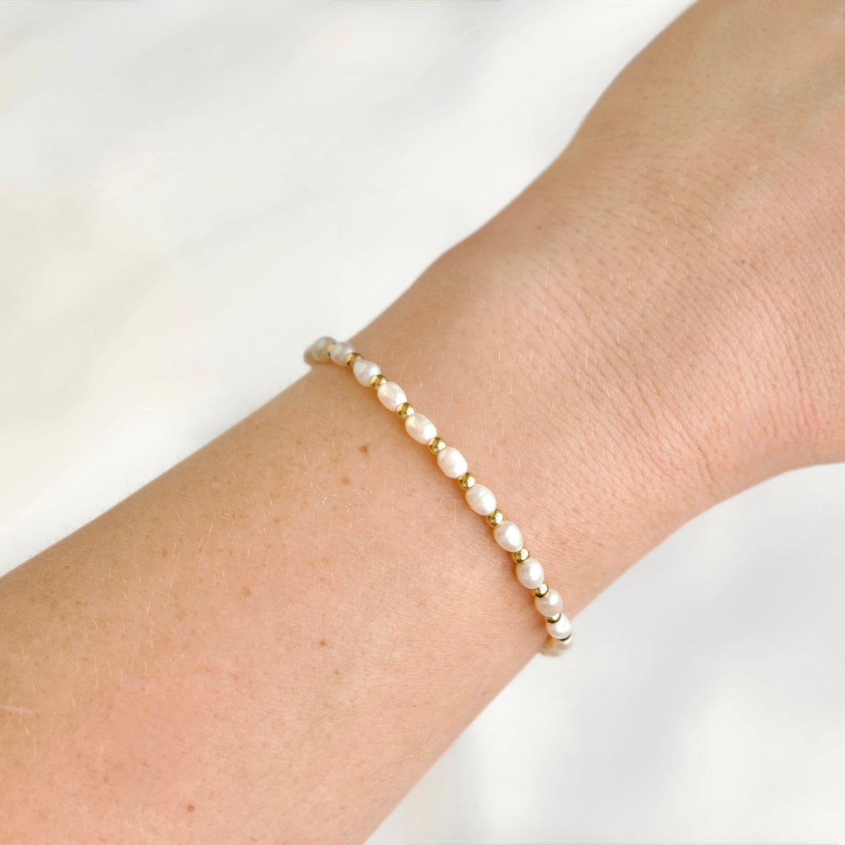 A delicate bracelet with small white pearls and gold spacer beads worn on a person's wrist against a soft white background.