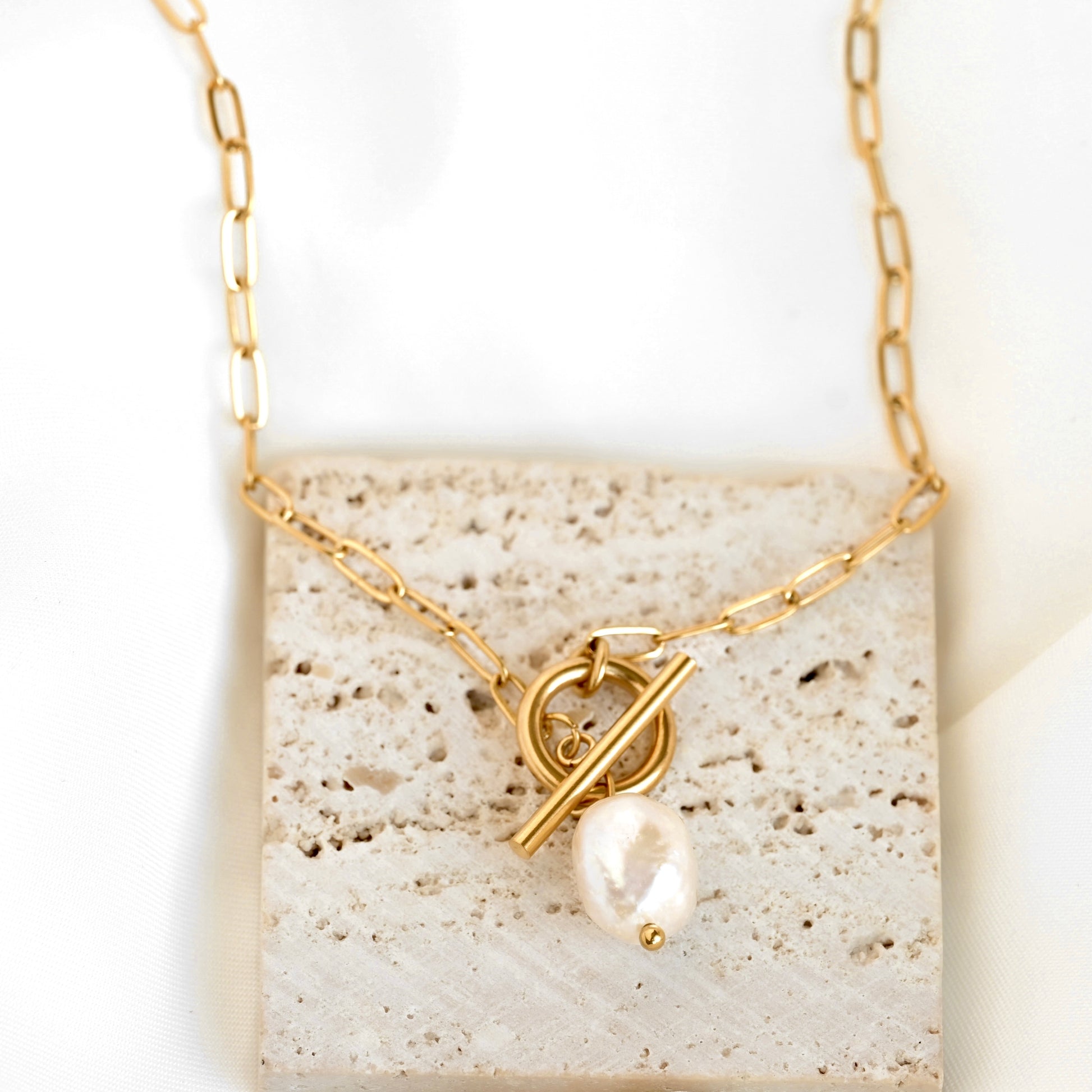 A gold chain necklace with a toggle clasp and a dangling baroque pearl, displayed on a textured beige stone tile against a white background.