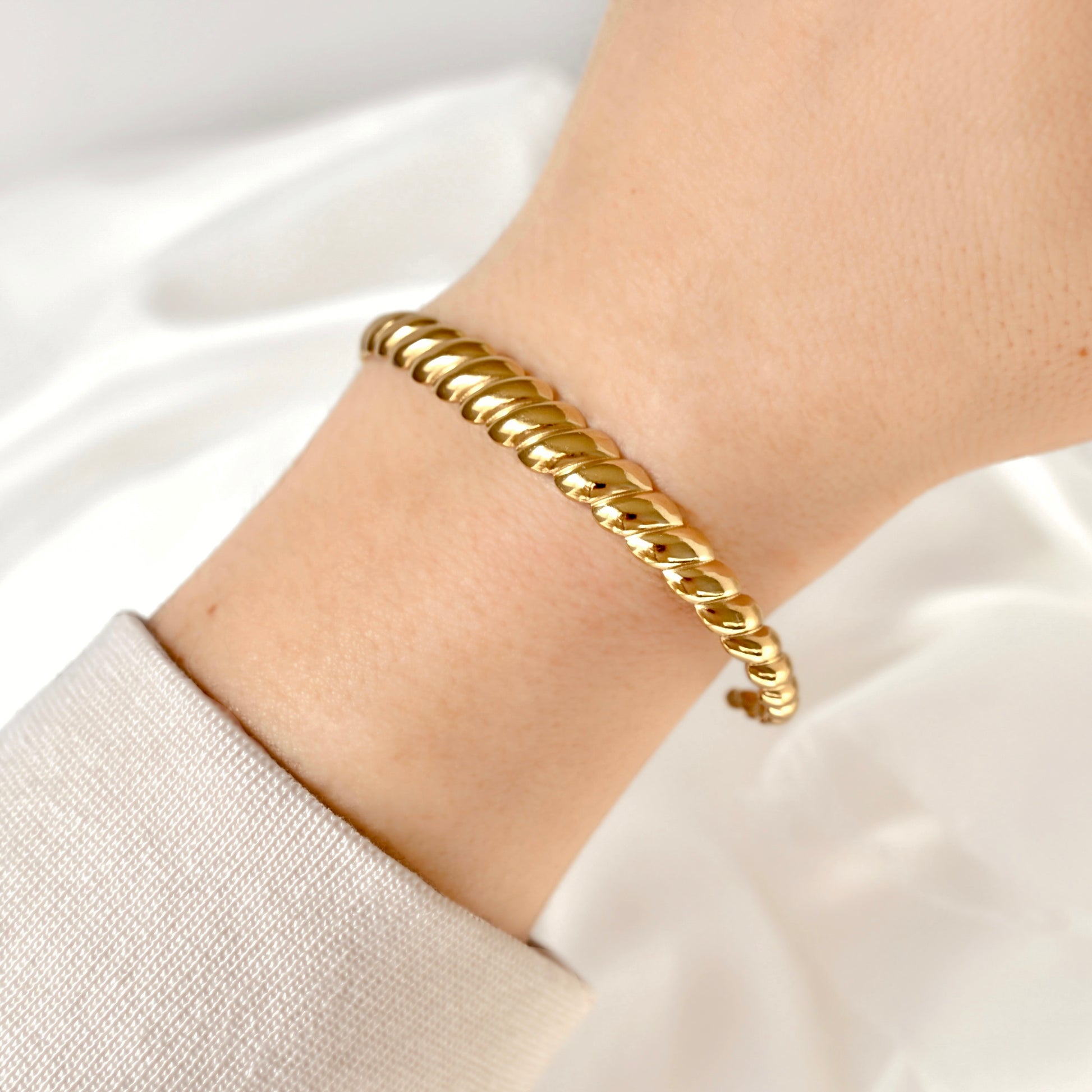 A close-up of a gold rope-style bangle bracelet worn on a person's wrist, with a light beige sleeve visible at the cuff against a soft white fabric background.