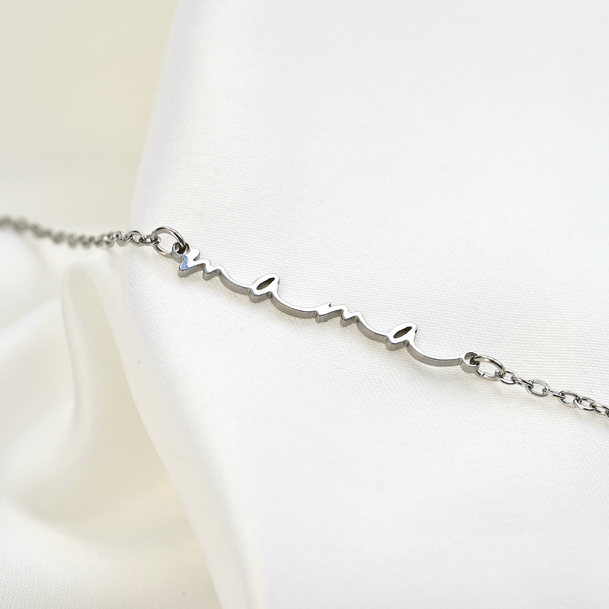 A silver personalized bracelet with a cursive script design reading 'mama', displayed on a white satin fabric background.