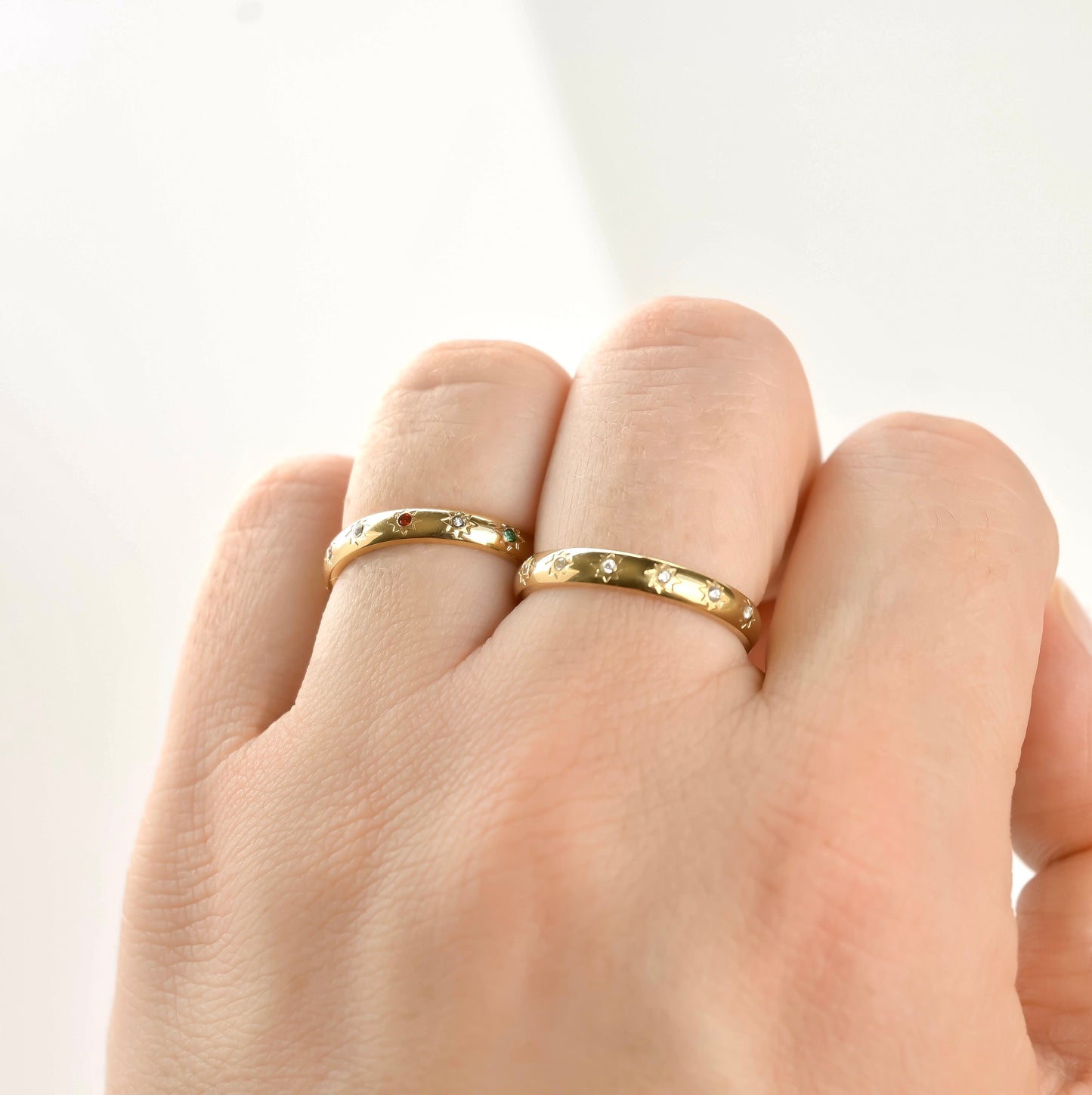 Two Gold rings, one with mixed color stones and one with clear stones  on a finger against a light background