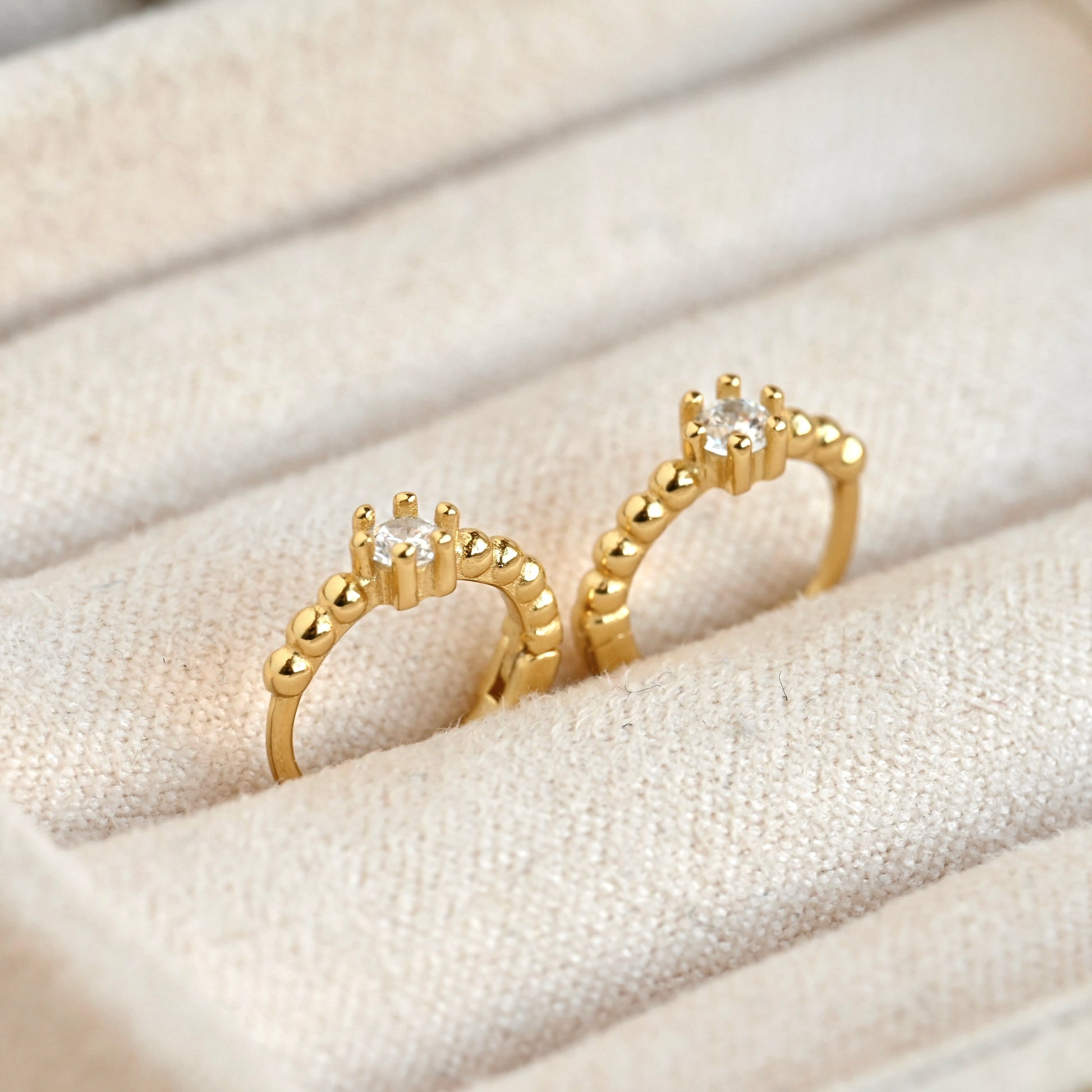 A pair of beaded gold hoop earrings with prong-set clear round gemstones, displayed on a textured beige fabric ring box.