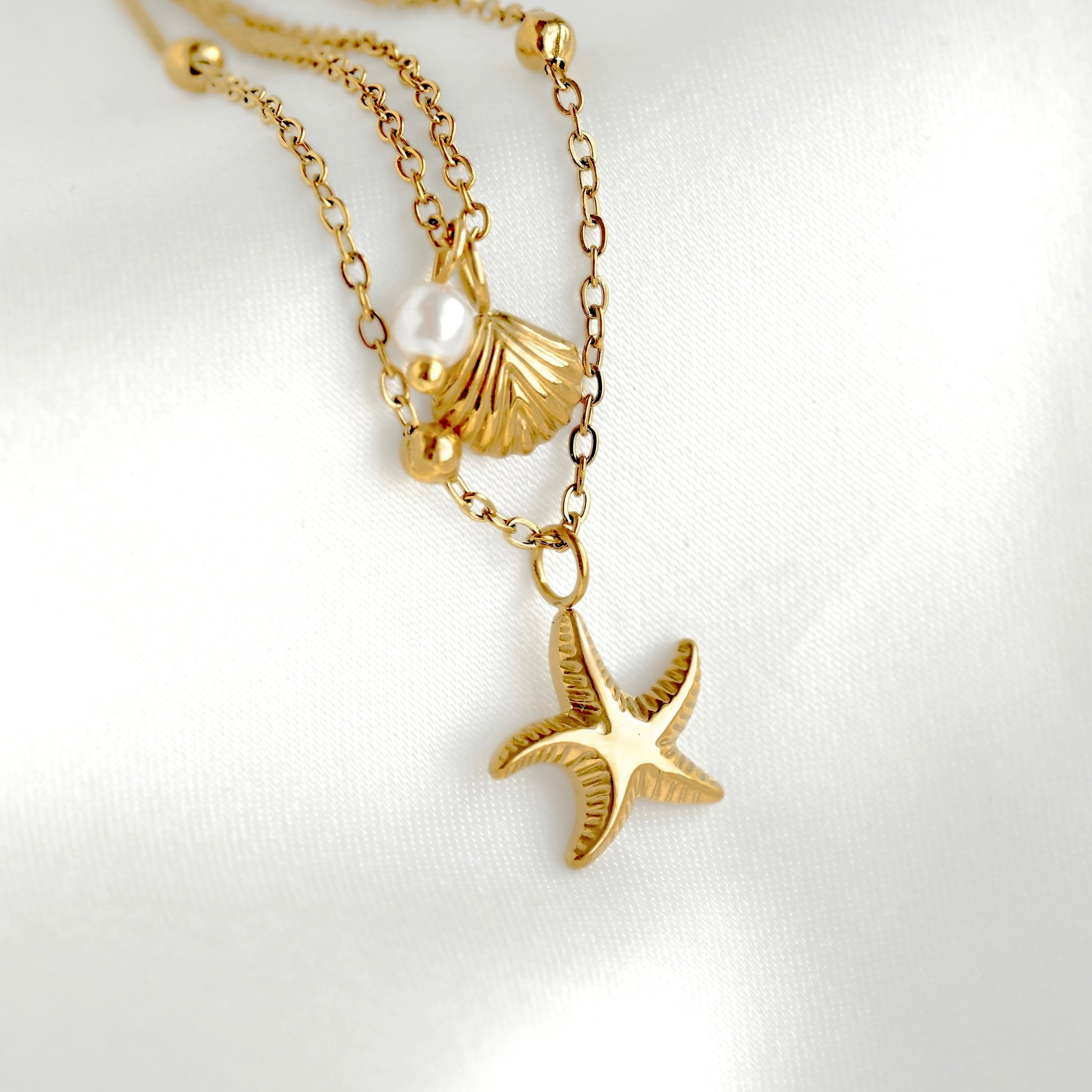A gold-toned layered necklace featuring a starfish pendant, a seashell charm, and a pearl bead, displayed on a soft white fabric background.