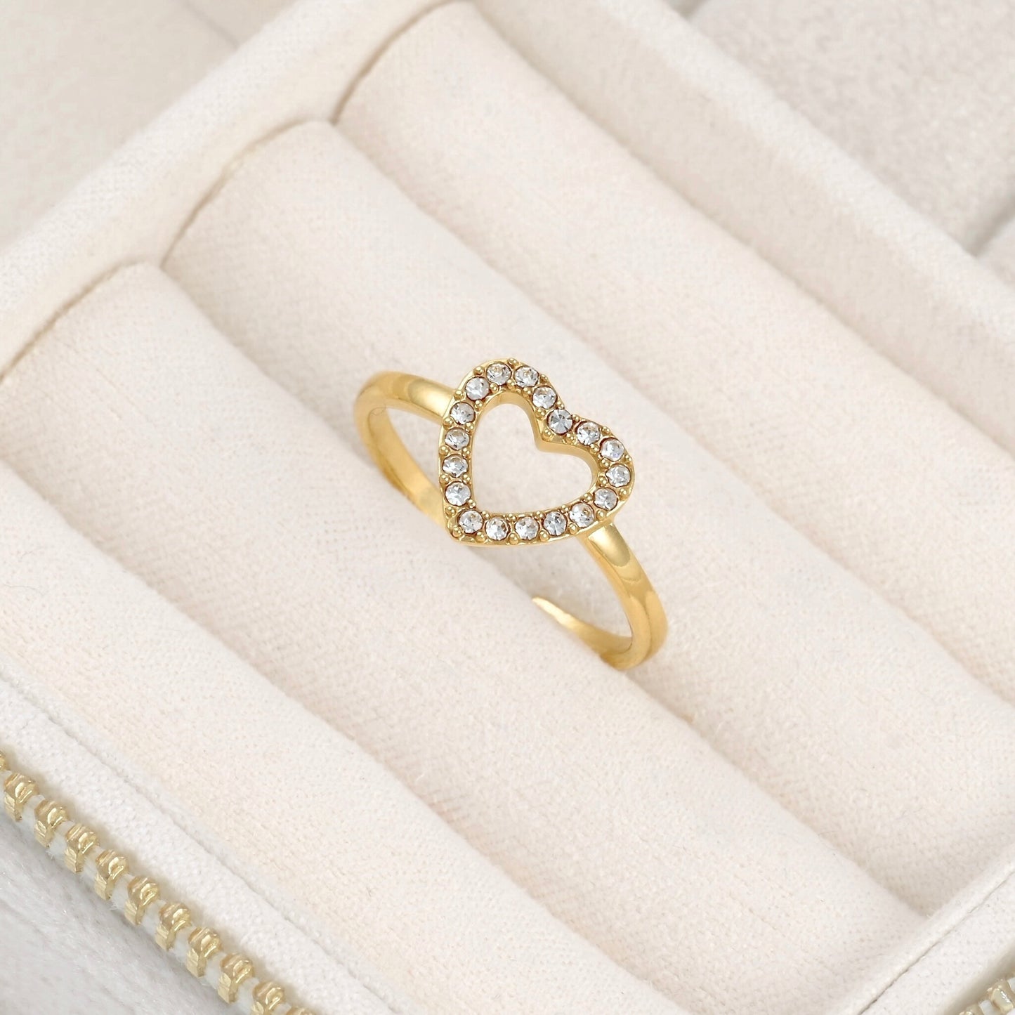 Top view of a gold ring featuring an open heart-shaped top set with small clear stones, displayed on a textured cream fabric surface.