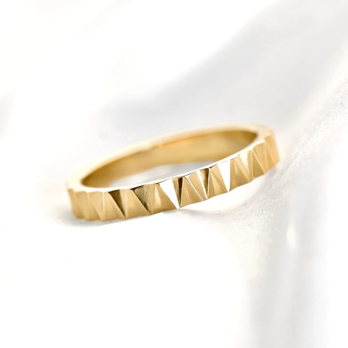 A gold ring with a textured, faceted triangular pyramid pattern around the outer band, resting on a soft white fabric background.
