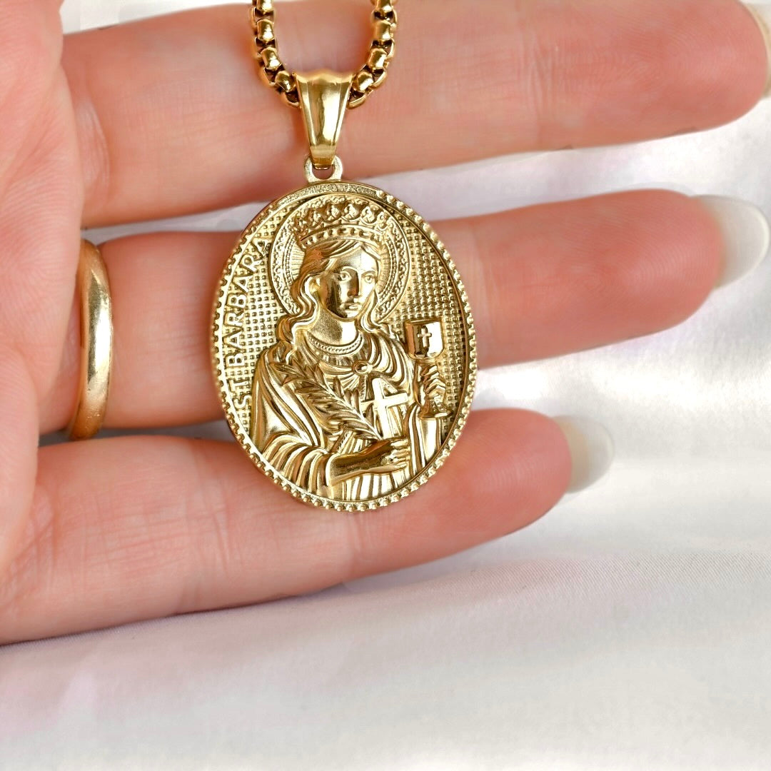 A gold oval religious pendant necklace featuring an embossed figure, held in a person's hand against a white fabric background.