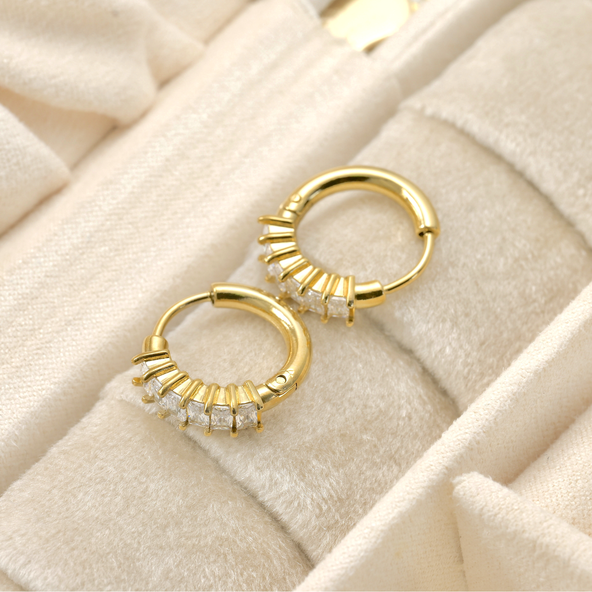Top view of small gold tube hoop earrings with embedded  rectangular CZ stones on a beige textured surface