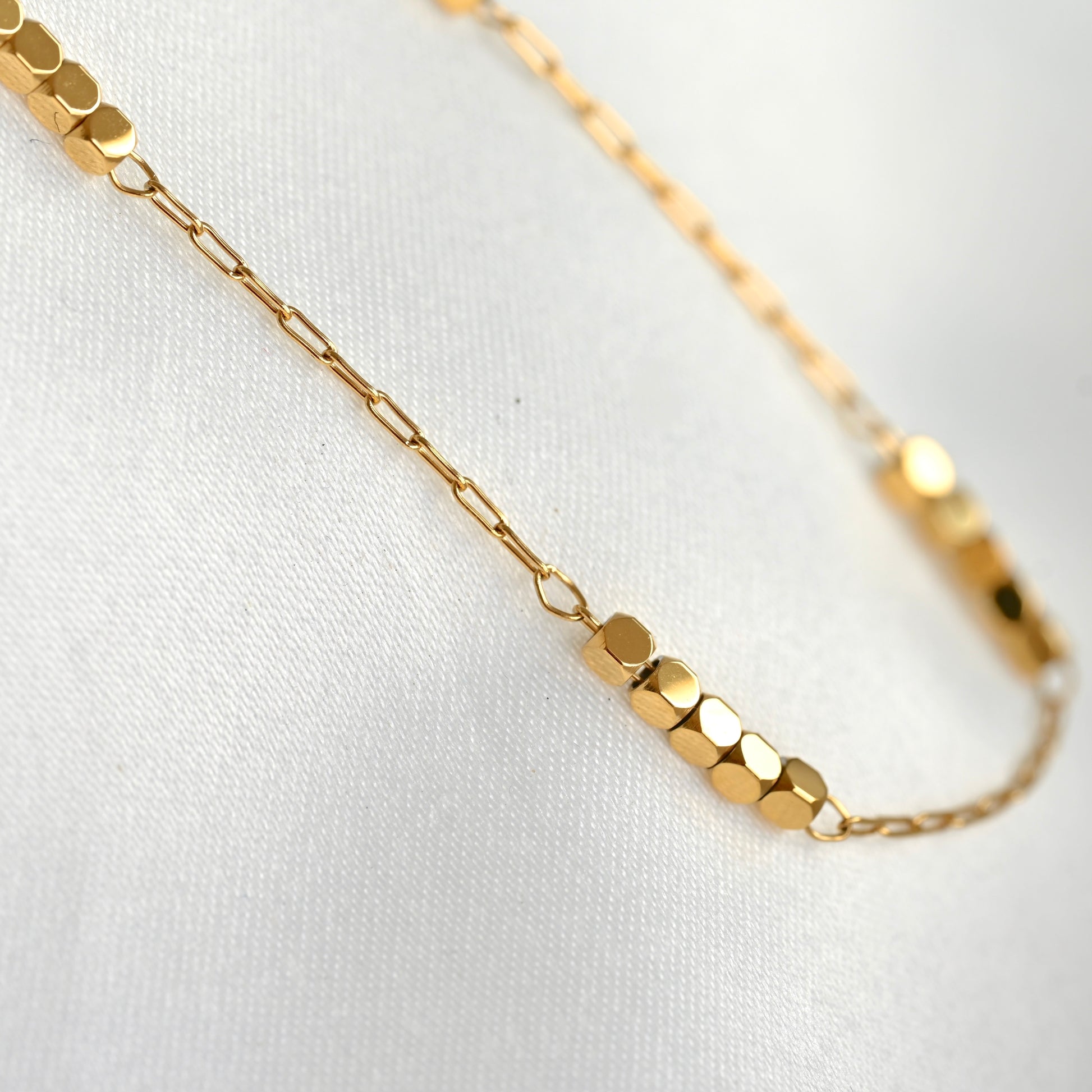 A delicate gold chain necklace featuring a central cluster of faceted hexagonal beads, displayed on a soft white textured fabric background.
