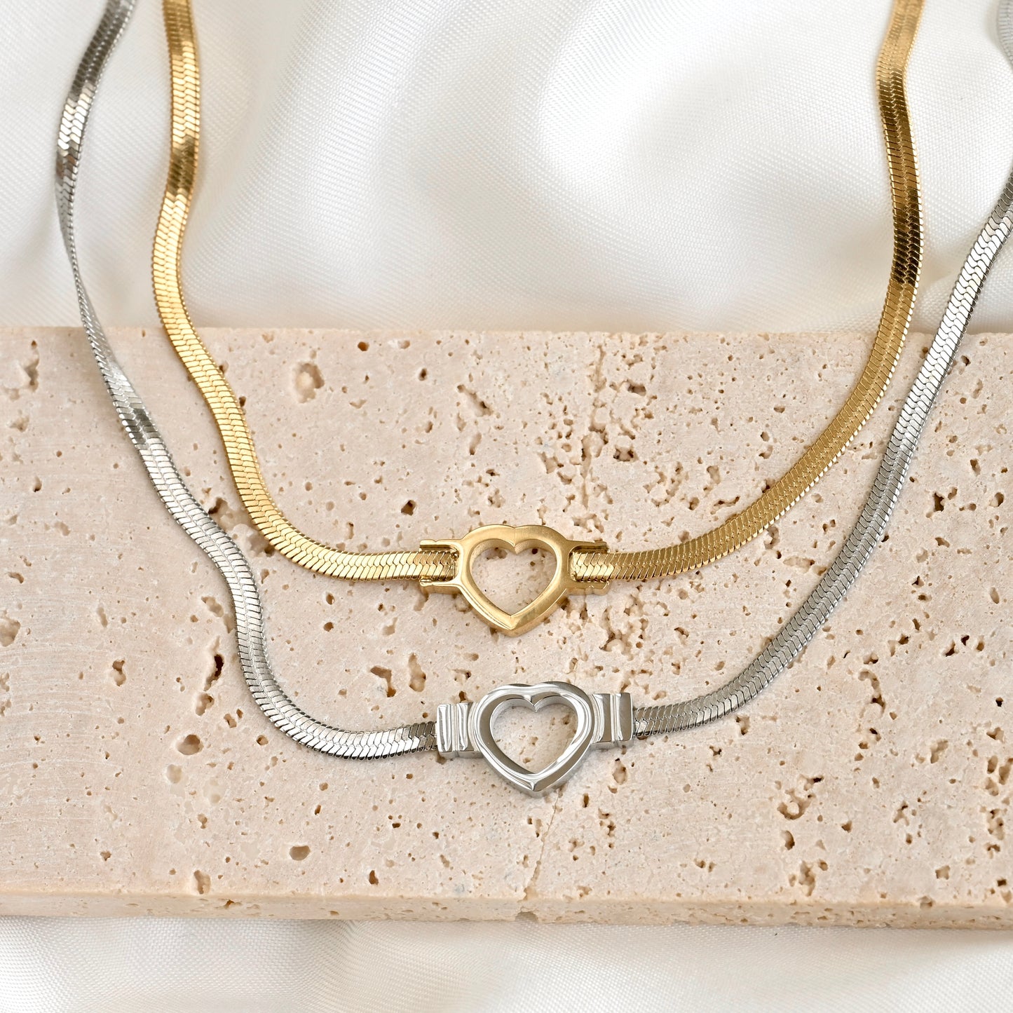 Two snake chain necklaces, one gold and one silver, with a cutout heart charm on a textured surface.