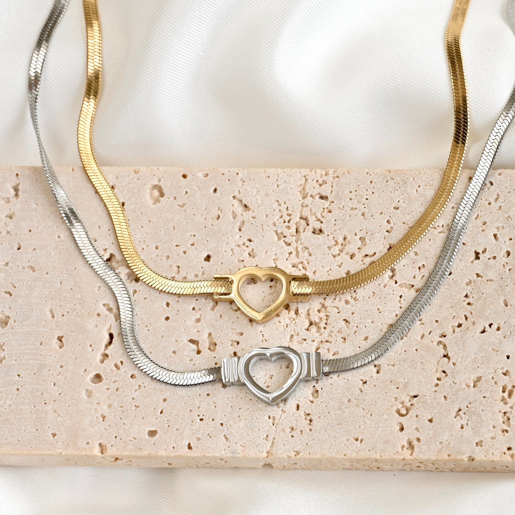 Two snake chain necklaces, one gold and one silver, with a cutout heart charm on a textured surface.