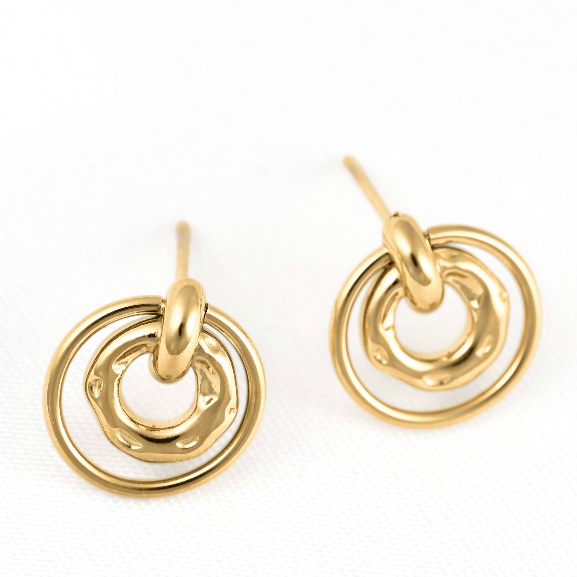 A pair of gold-toned dangle earrings with a textured, hammered finish and concentric circular designs, displayed on a clean white textured background.