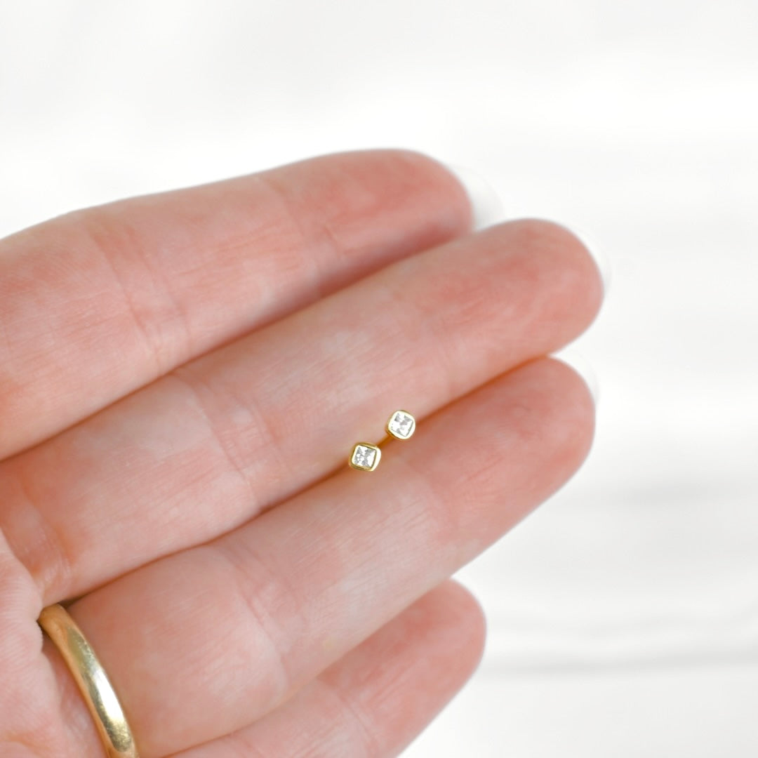 A close-up of a person's hand holding two tiny gold initial stud earrings, with a gold band ring visible on the ring finger against a soft white background.