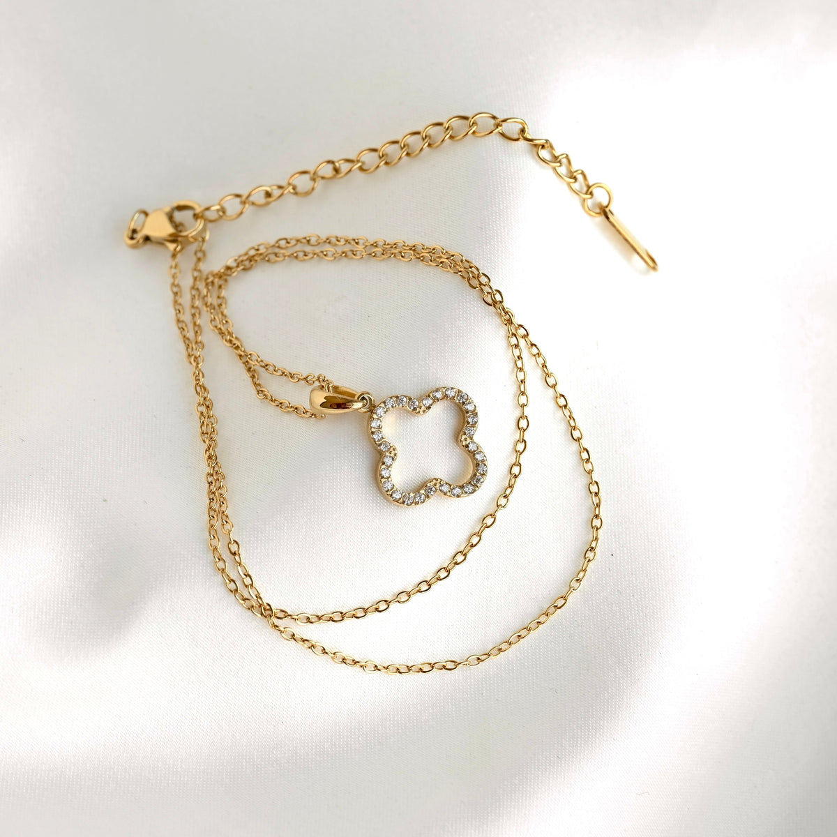 A gold-toned chain necklace with a clover-shaped pendant set with clear crystals, displayed on a white textured fabric background.