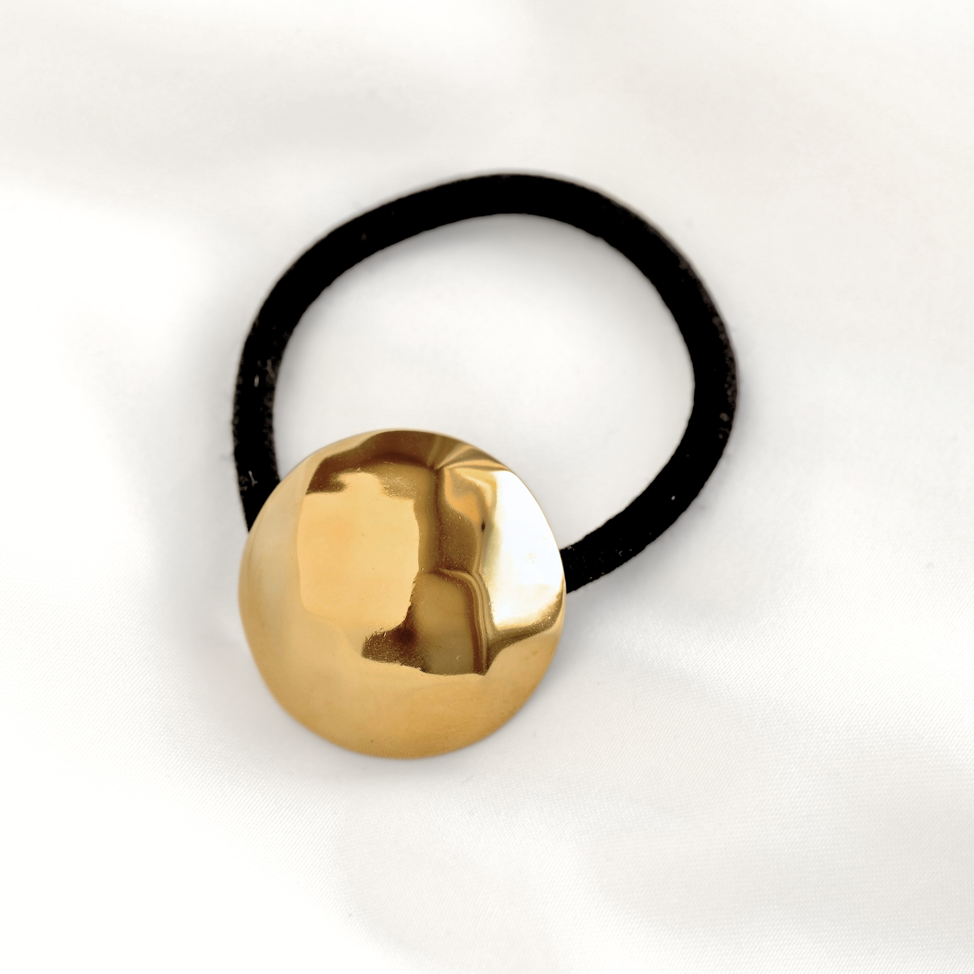 A black elastic hair tie with a large, round, polished gold disc pendant, photographed on a white fabric background.