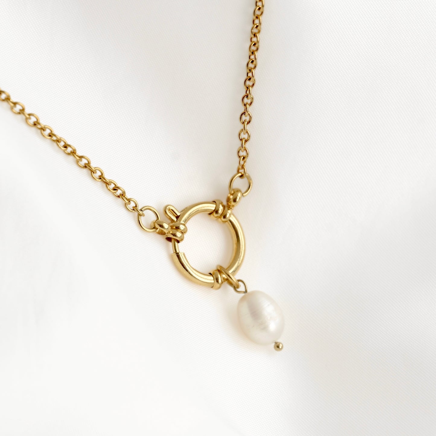 A gold chain necklace featuring a circular ring pendant with a small white pearl dangle, displayed on a soft white fabric background.