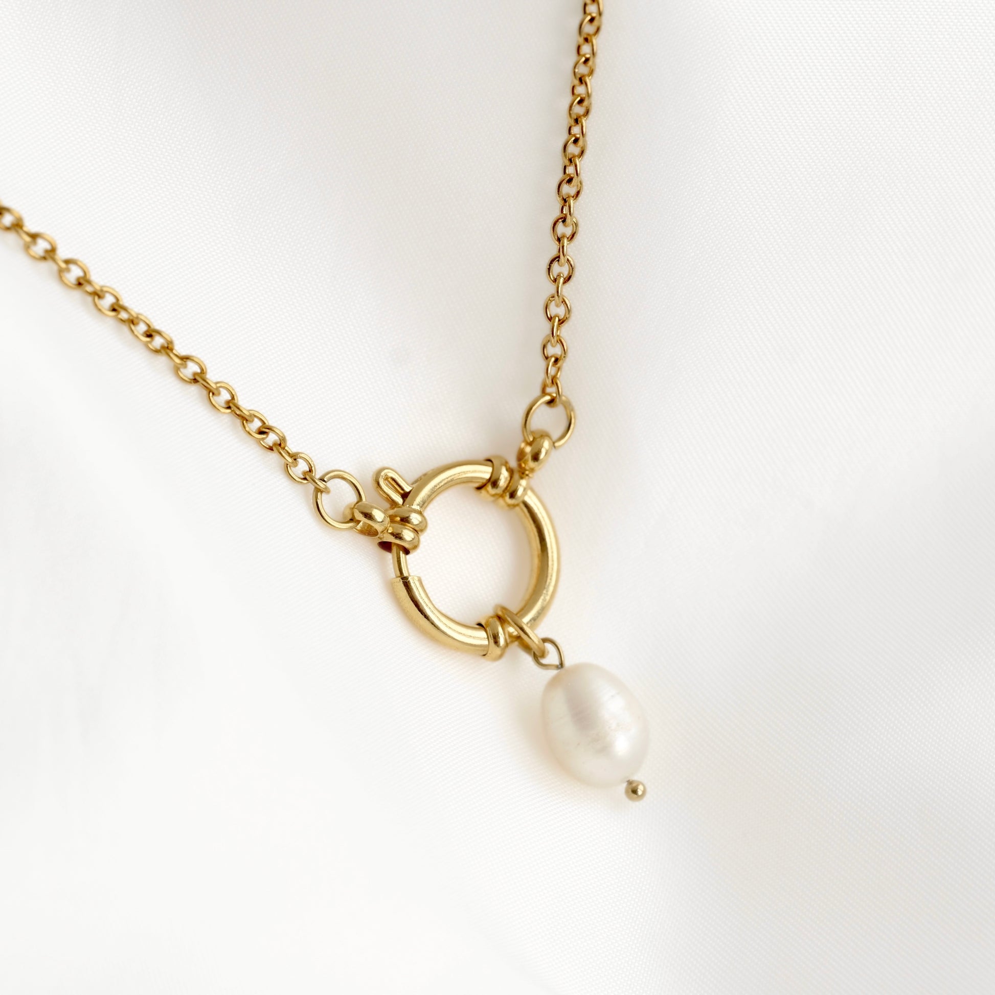 A gold chain necklace featuring a circular ring pendant with a small white pearl dangle, displayed on a soft white fabric background.
