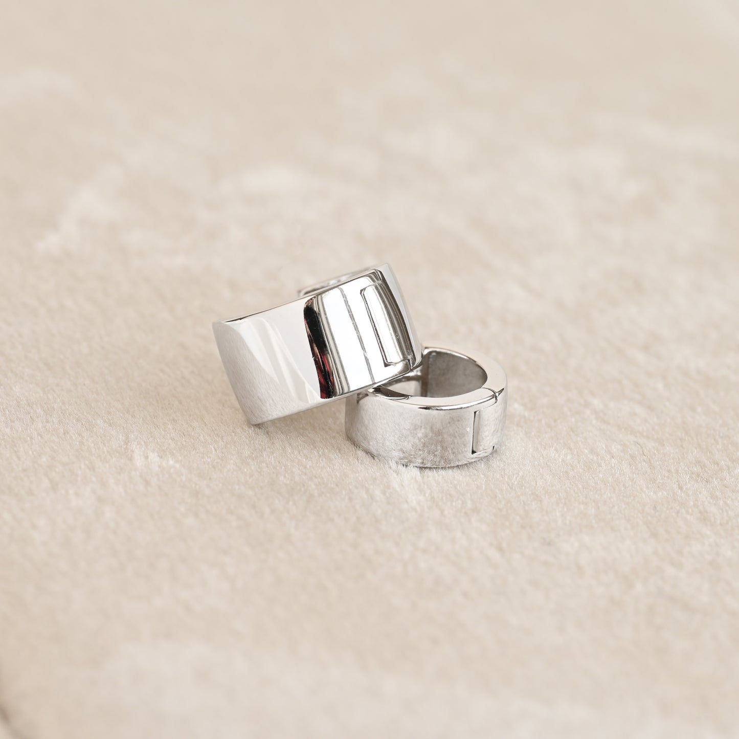 Two polished silver bold hinged hoop earrings with a modern, geometric block design, resting on a soft beige textured surface.