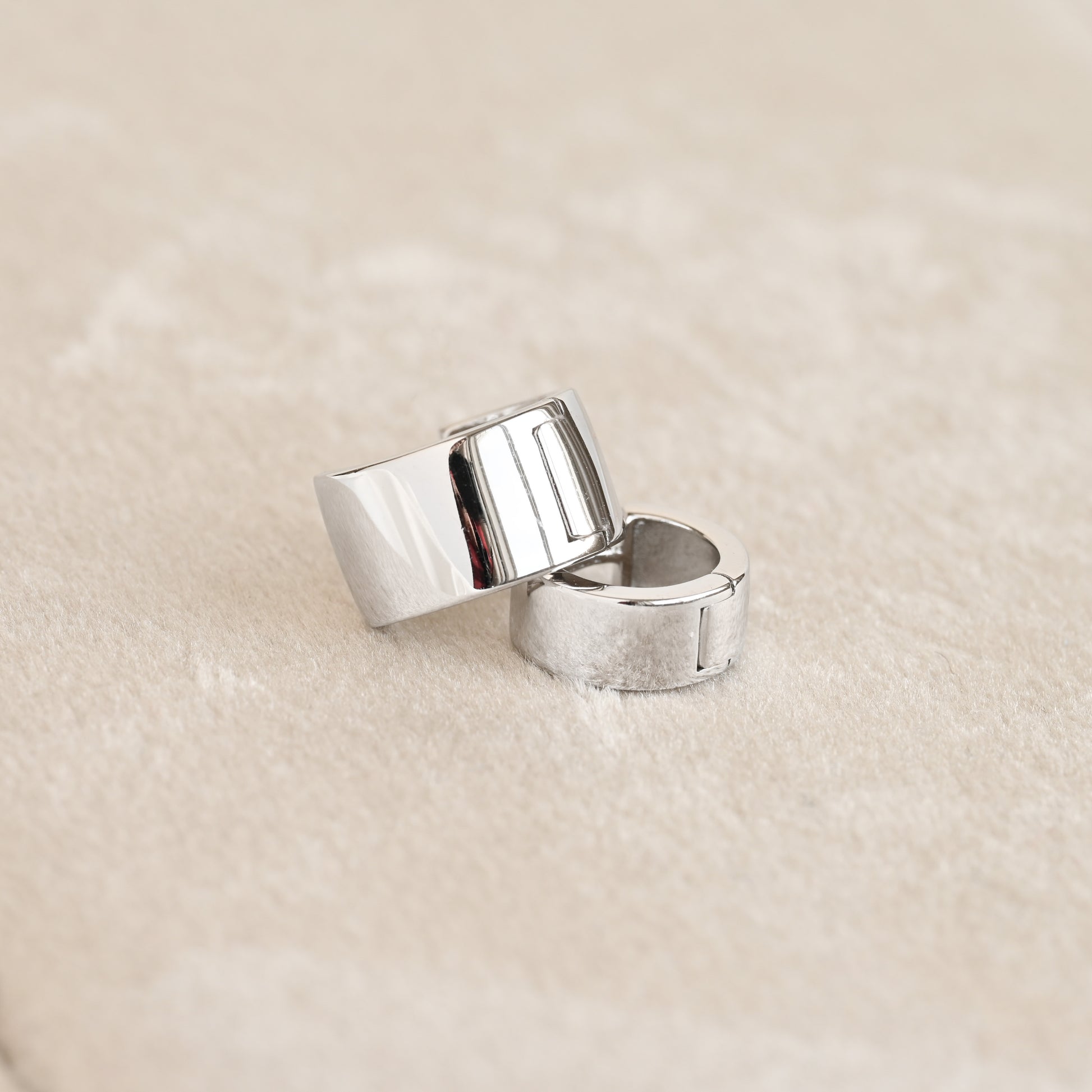 Two polished silver bold hinged hoop earrings with a modern, geometric block design, resting on a soft beige textured surface.