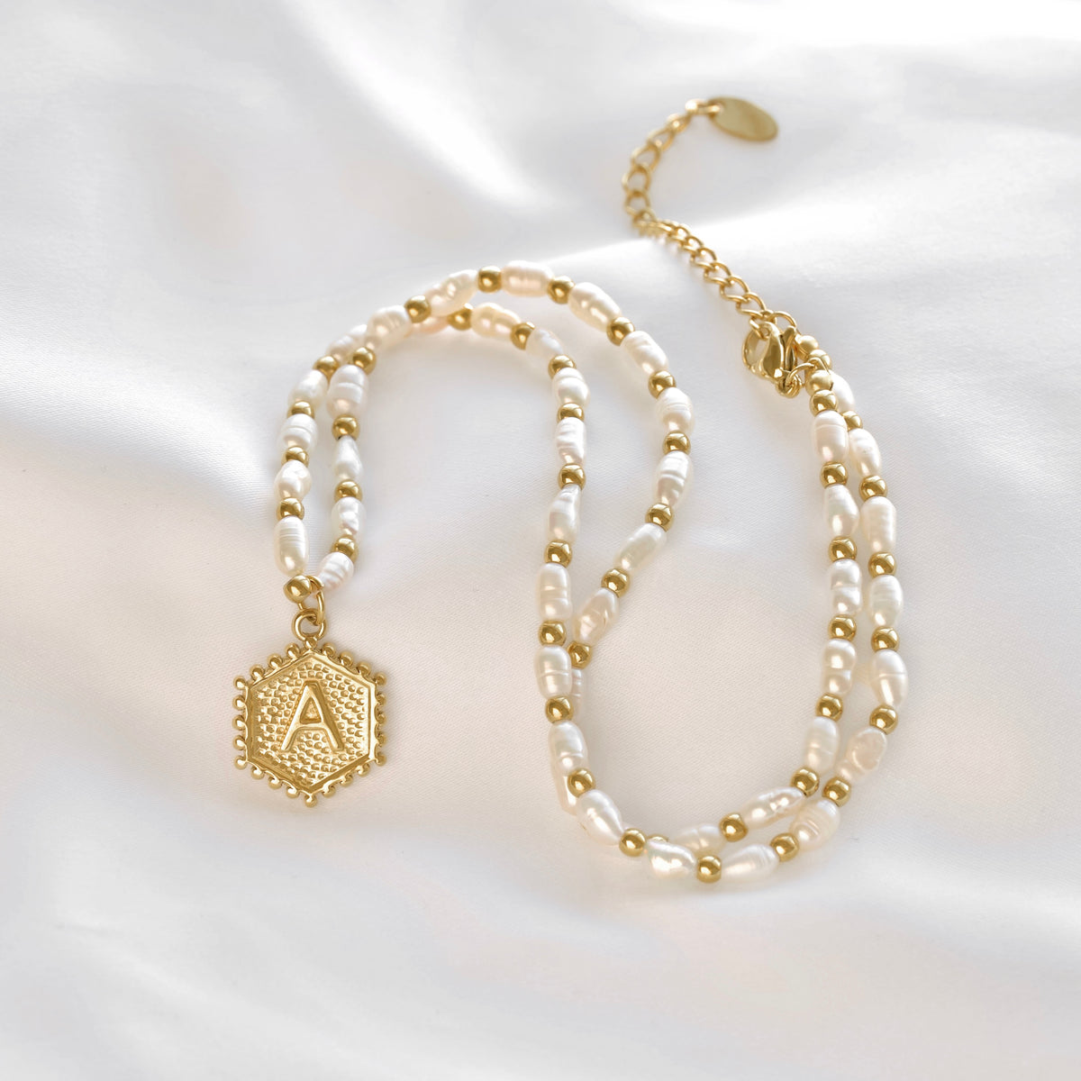 A double-strand pearl necklace with gold bead accents and a gold initial 'A' hexagonal pendant, displayed on a soft white satin fabric background.