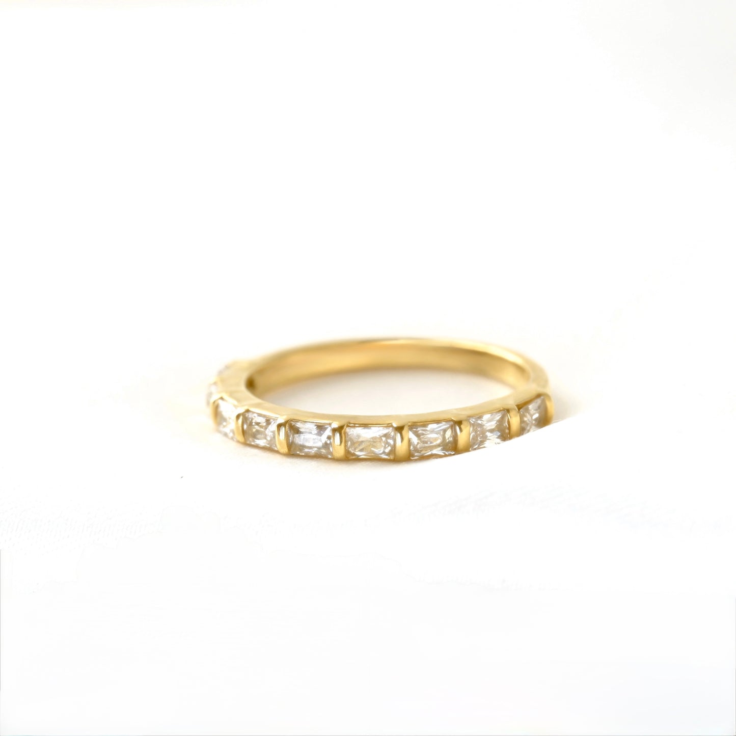 A yellow gold half eternity ring featuring a row of baguette-cut clear gemstones, photographed on a seamless white background.