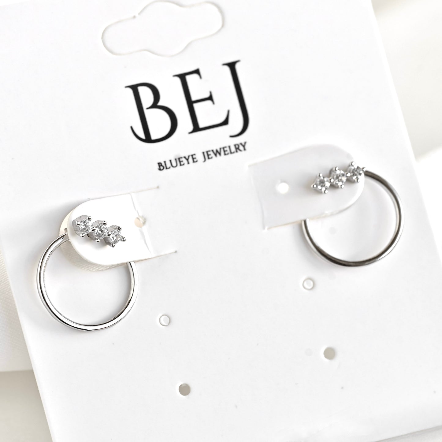 A pair of silver-toned  ear jacket earrings with small clear crystal clusters on the front and a polished circle at the back, displayed on a white card from Blueeye Jewelry. 