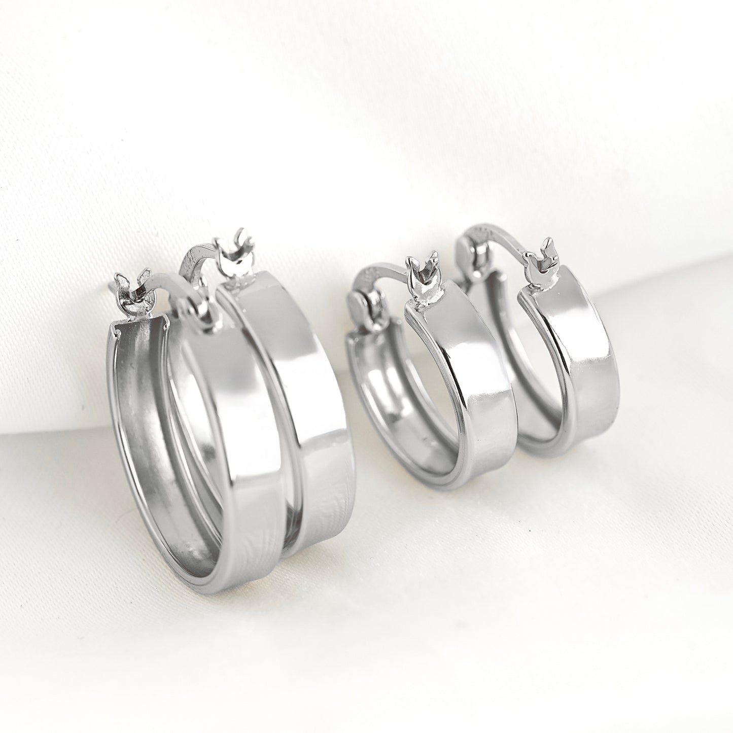A pair of polished silver-tone hoop earrings with latch back clasp, displayed on a soft white fabric background.