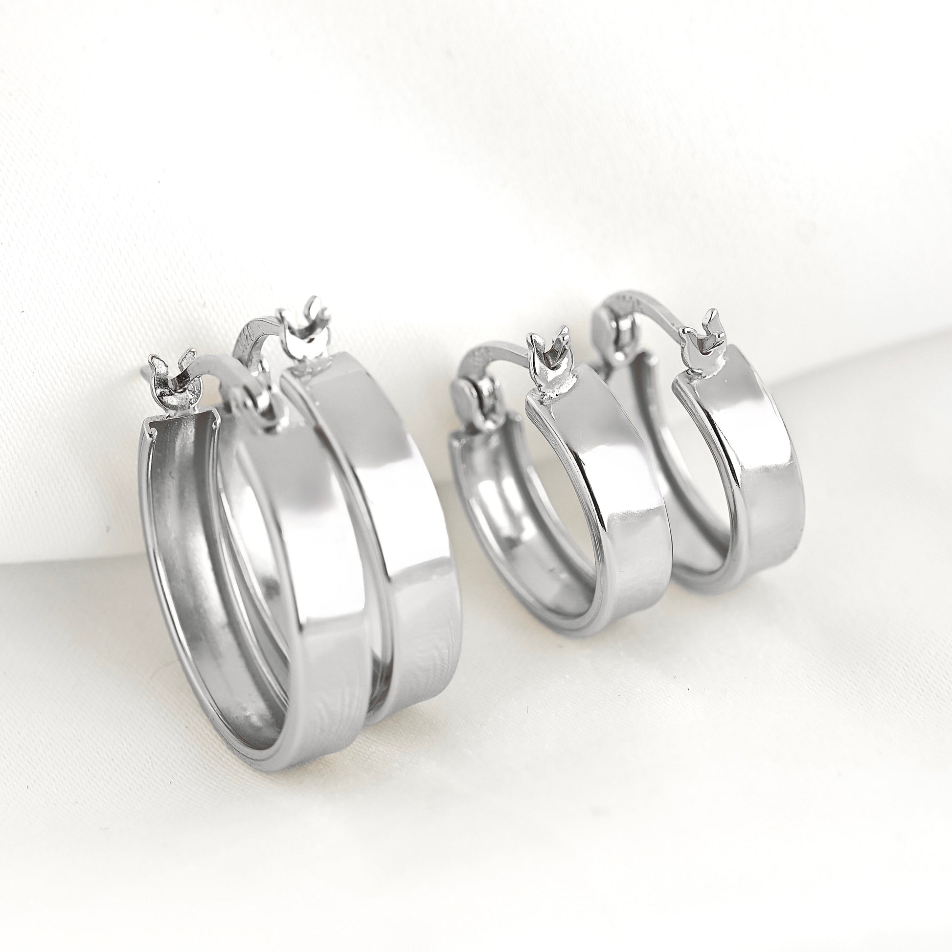A pair of polished silver-tone hoop earrings with latch back clasp, displayed on a soft white fabric background.