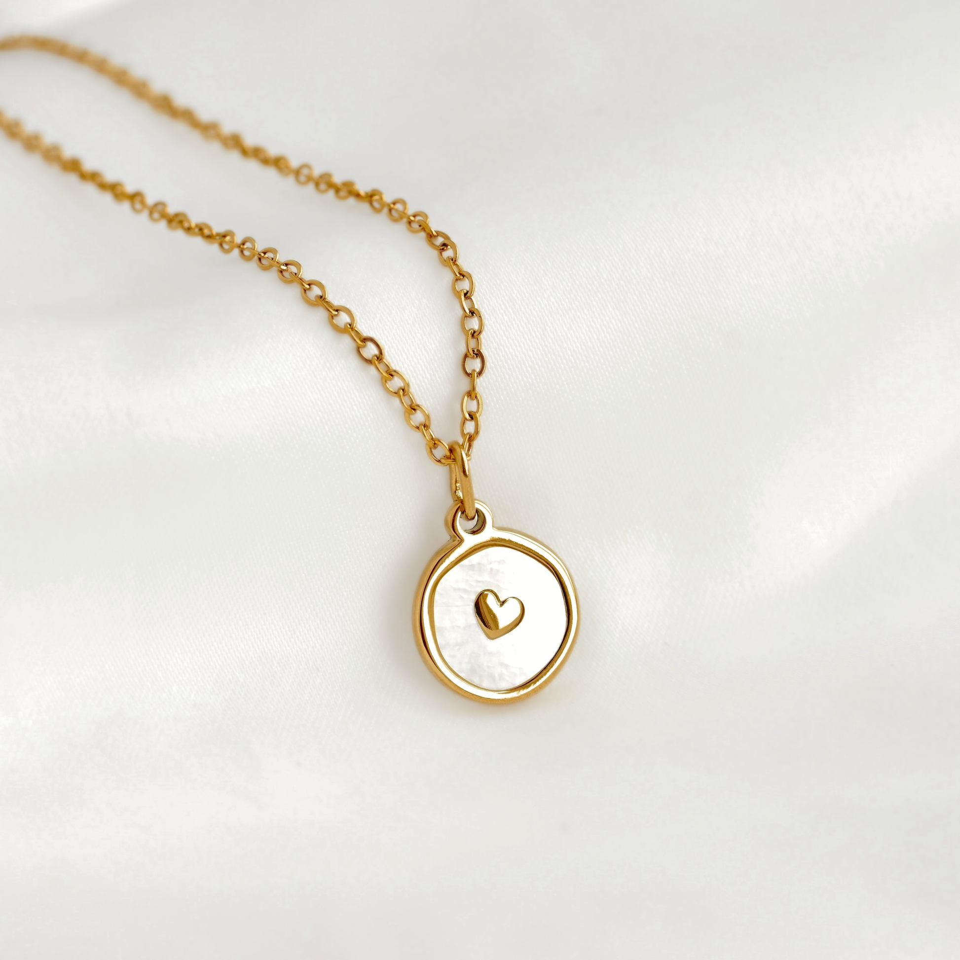 A delicate gold chain necklace with a round pendant featuring a white inlay and a small gold heart in the center, displayed on a soft white fabric background.