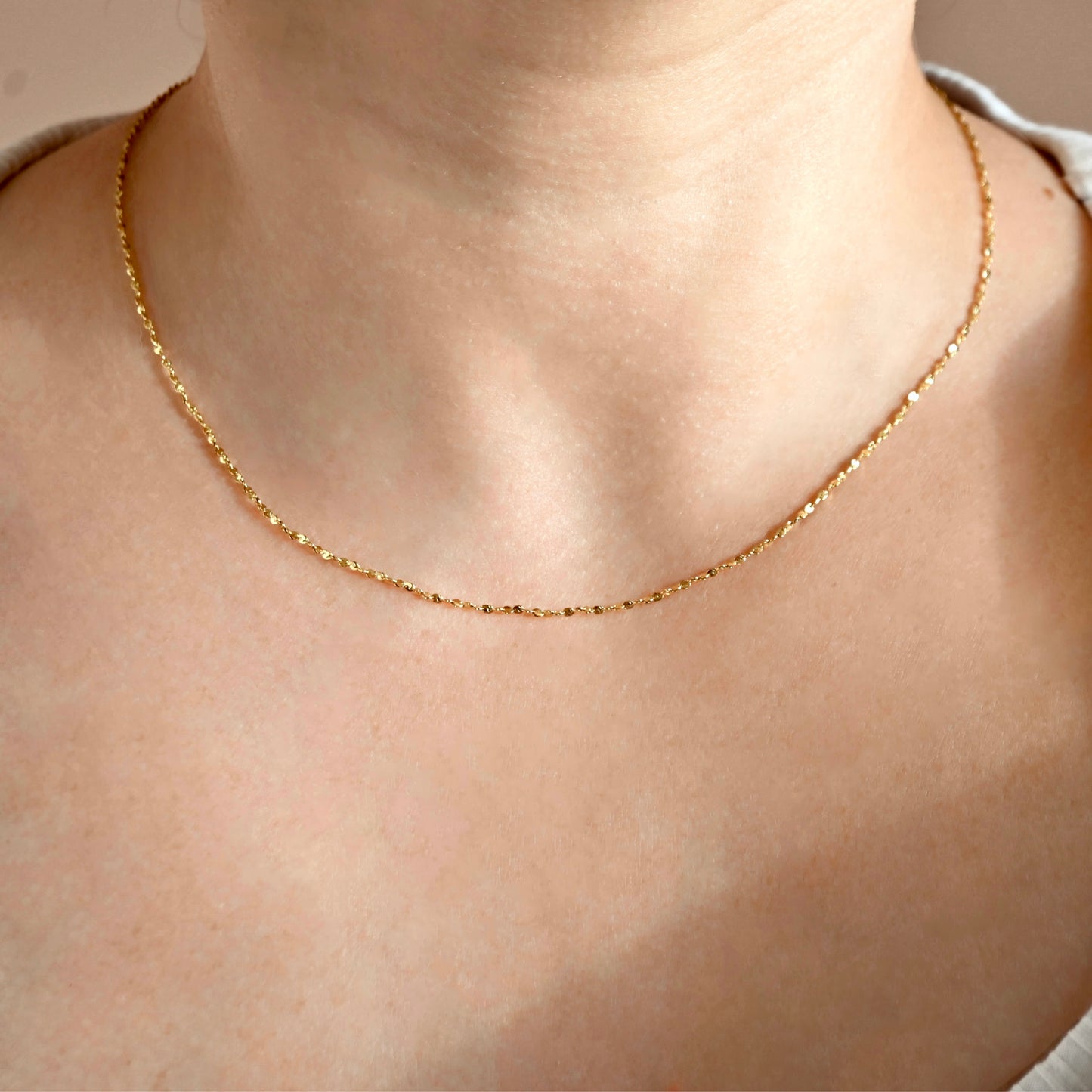 A delicate gold chain necklace worn on a person's neck, shown in a close-up shot against a soft beige background.