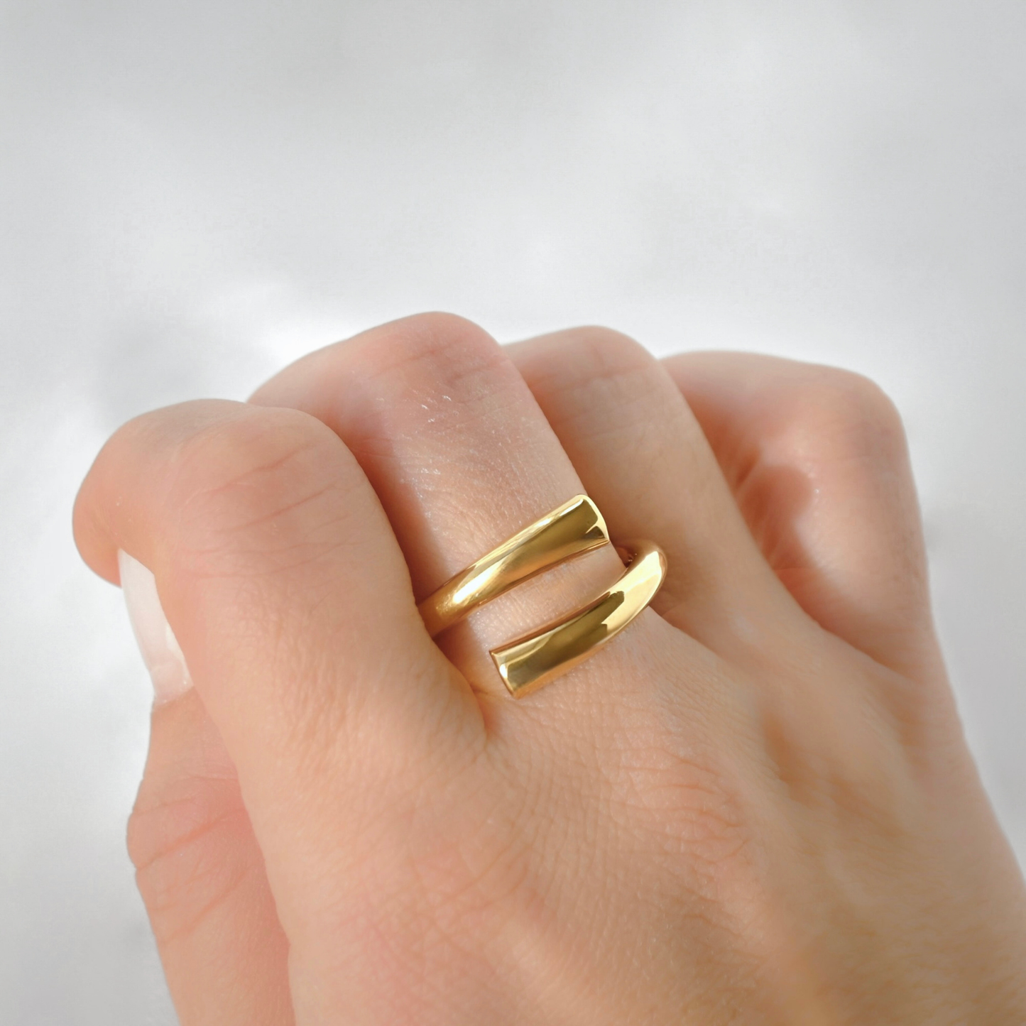 A close-up of a hand wearing a chunky gold ring with two parallel polished bands, photographed against a soft light gray background.
