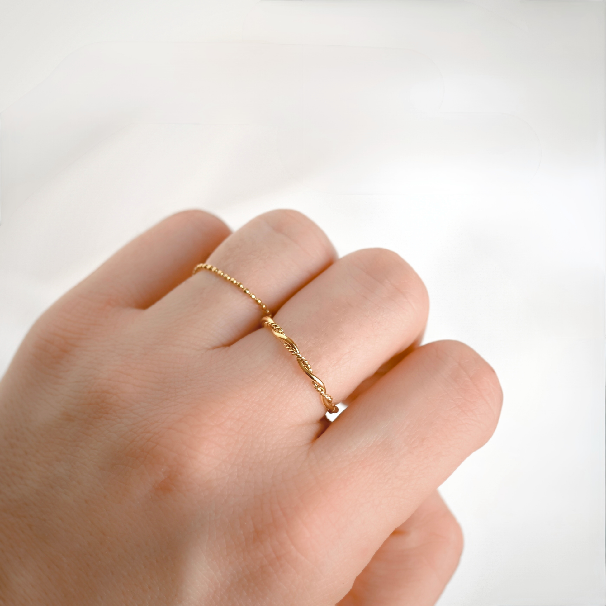A delicate gold chain ring on ring finger and a entwined rope ring on middle finger, shown worn on a person's finger against a soft white background.