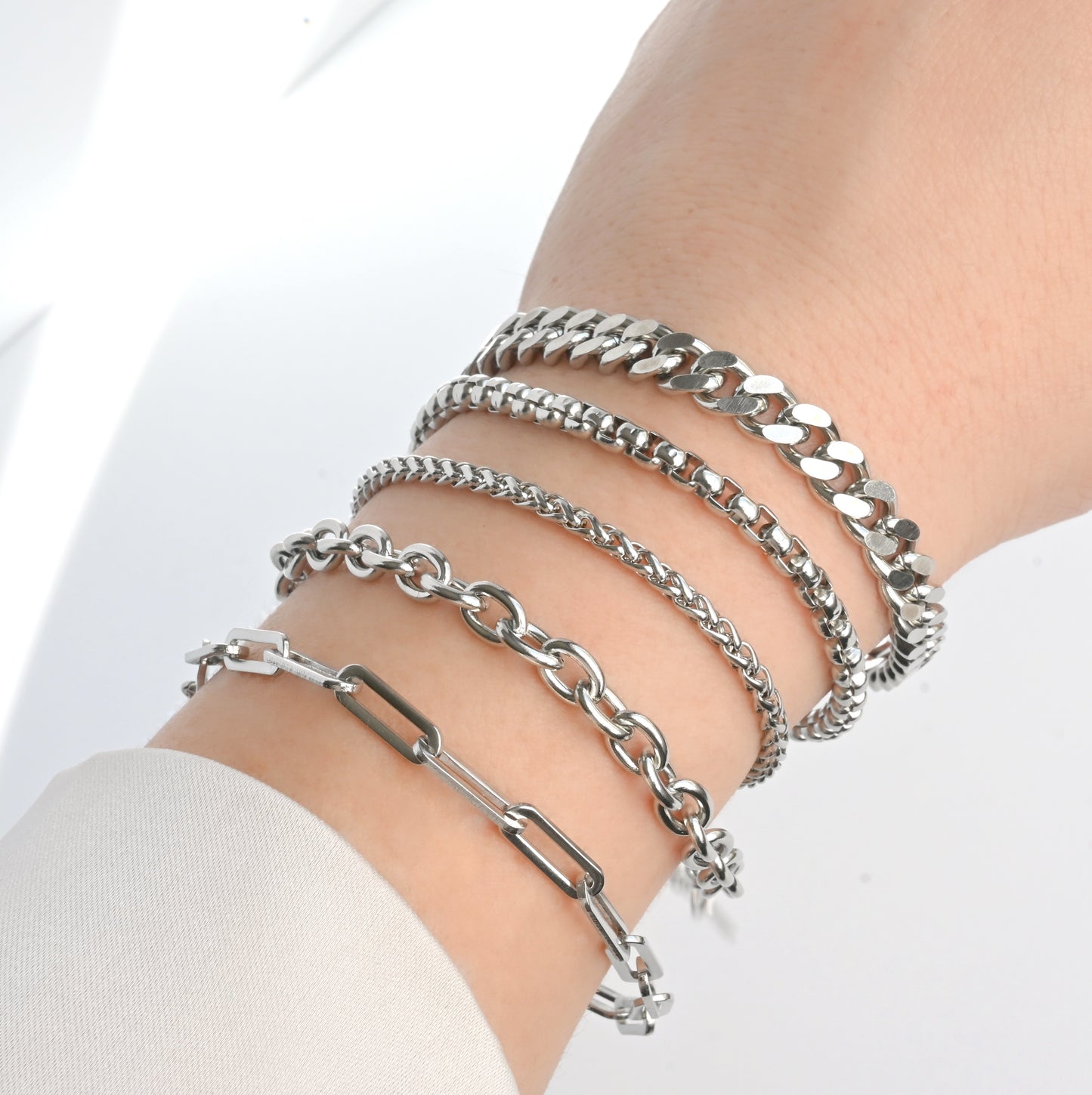 Silver chain bracelets (bold paperclip, rolo, wheat, box and cuban chains) on a person's wrist against a white background