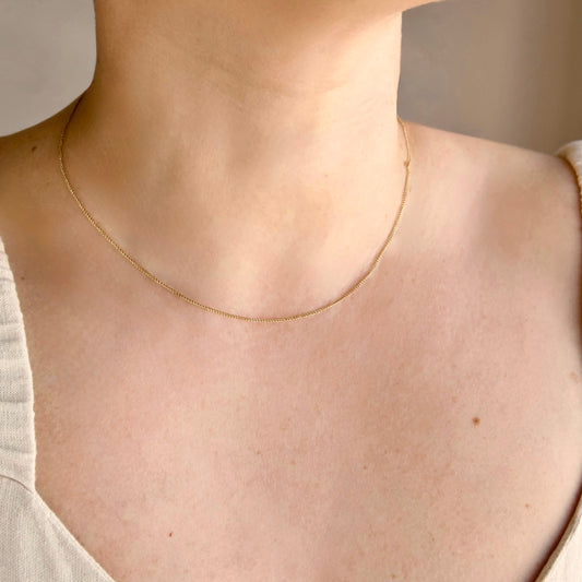An ultra fine gold curb chain necklace worn on a person's neck, shown in a close-up shot against a soft neutral background.